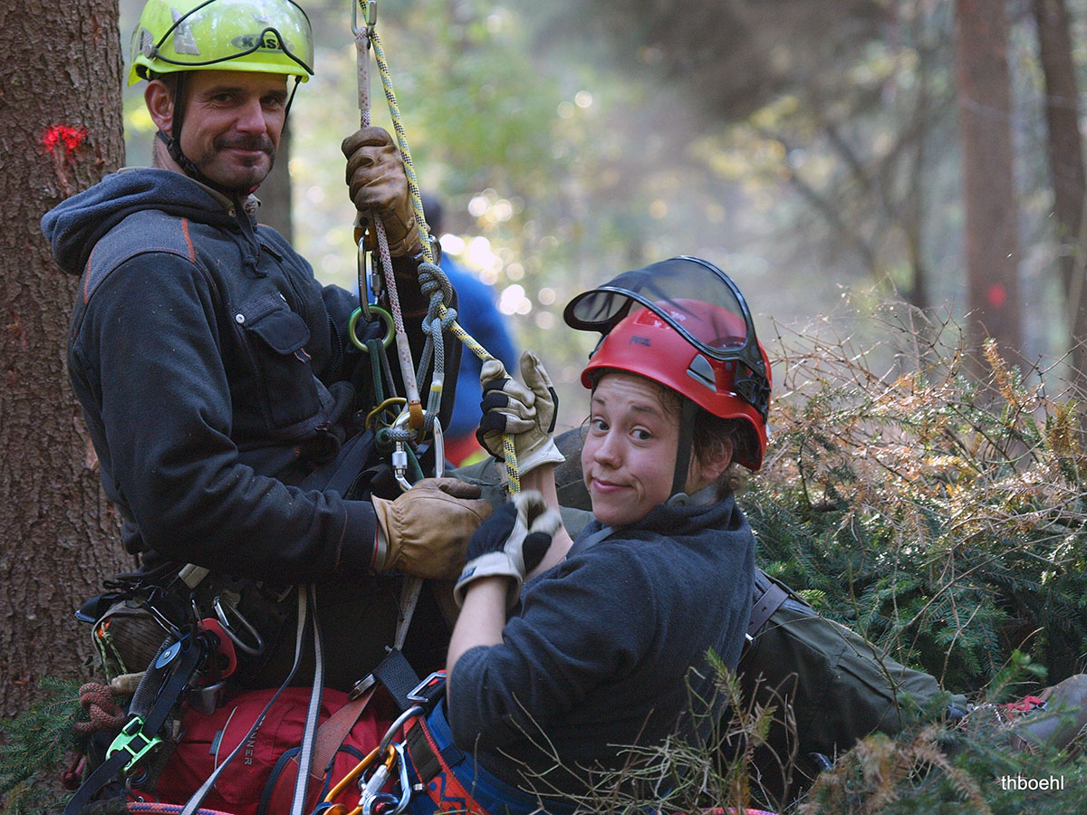 Zwei Baumpfleger mit Helmen und Klettergurten lächeln in die Kamera, während sie in einem Wald mit Seilen arbeiten. Sie zeigen ihre Seilkletterausbildung, während sie sich beim Klettern oder bei Wartungsarbeiten geschickt durch die Bäume und Äste bewegen.