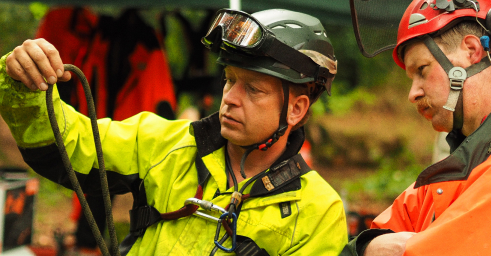 Zwei Männer in Sicherheitsausrüstung untersuchen ein Seil im Freien während einer Seilkletterausbildung. Der eine trägt einen grünen Helm und eine gelbe Jacke und hält das Seil, während der andere mit rotem Helm und oranger Jacke aufmerksam zusieht.