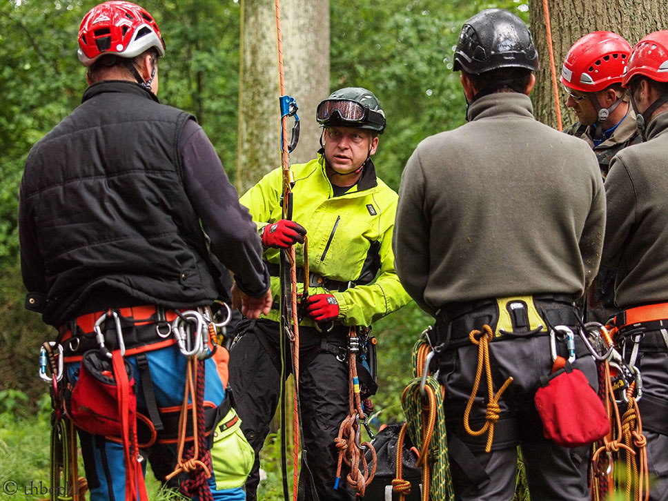 Eine Gruppe von Personen mit Helmen und Sicherheitsausrüstung steht in einem Wald und hört einem Ausbilder in einer leuchtend gelben Jacke zu, der ein Seil hält und Kletter- oder Rettungstechniken erklärt.