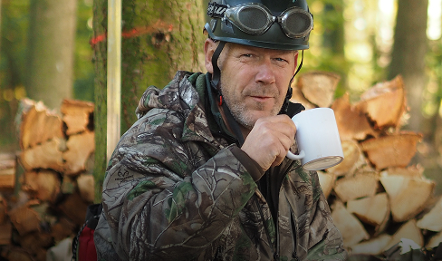 Ein Mann in Tarnkleidung und mit Schutzbrille nippt in einem Waldgebiet an einem weißen Becher, hinter ihm liegt ein Stapel gehacktes Brennholz.