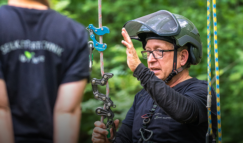 Eine Person, die einen Helm und eine Brille trägt, erklärt mit Gesten eine an Seilen befestigte Kletterausrüstung im Freien, während eine andere Person in der Nähe steht. Im Hintergrund ist grünes Laub zu sehen.