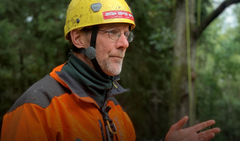 Ein Mann mit einer orangefarbenen Jacke und einem gelben Helm steht im Freien, trägt eine Brille und gestikuliert mit den Händen. Grüne Bäume sind im Hintergrund verschwommen.