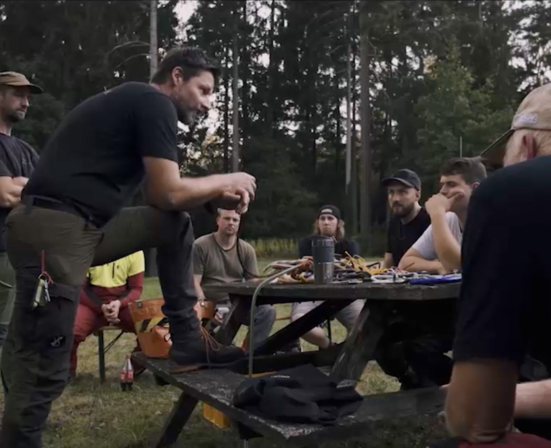 Eine Gruppe von Menschen sitzt und steht um einen hölzernen Picknicktisch im Freien in einem Waldgebiet und hört aufmerksam einem Mann zu, der über Seilkletterausbildung spricht. Auf dem Tisch stehen verschiedene Geräte und Getränke. Die Stimmung scheint ernst und konzentriert zu sein.